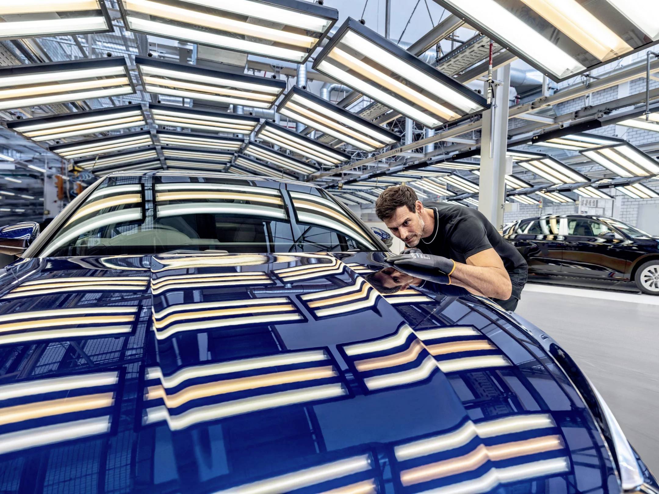 Ein Mann inspiziert sorgfältig die glänzende blaue Motorhaube eines Autos in einem gut beleuchteten Fabrikbereich.