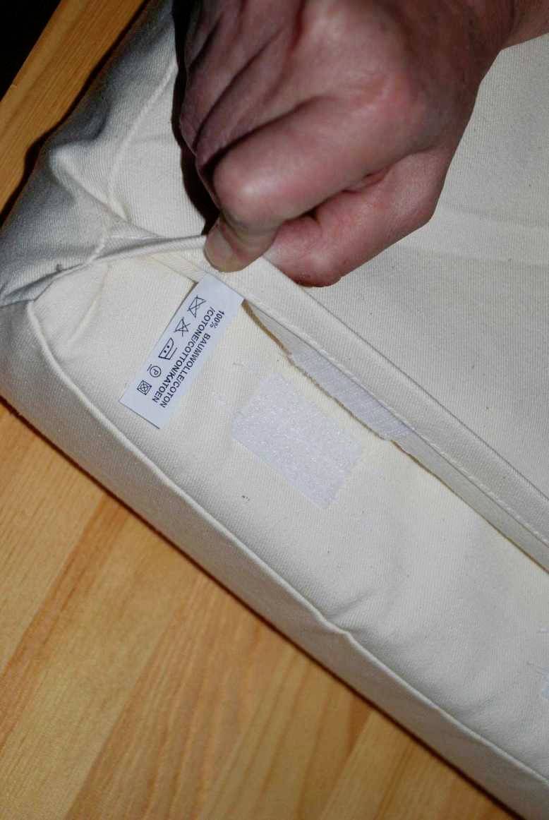 Eine Hand zieht an einem Etikett eines weißen Kissens, das Pflegehinweise und Symbole für Handwäsche und Bügeln zeigt.