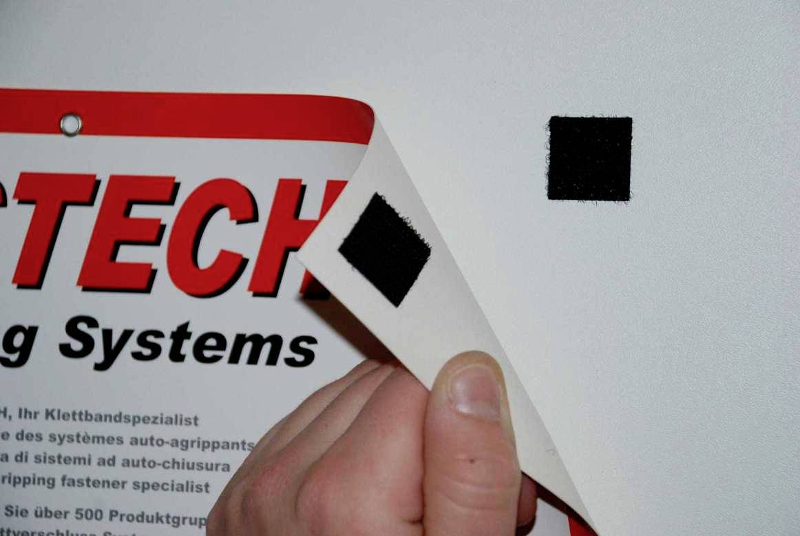 Eine Hand löst ein Stück Klettverschluss von einer weißen Oberfläche. Rechts daneben ist ein weiteres befestigtes schwarzes Klettstück.