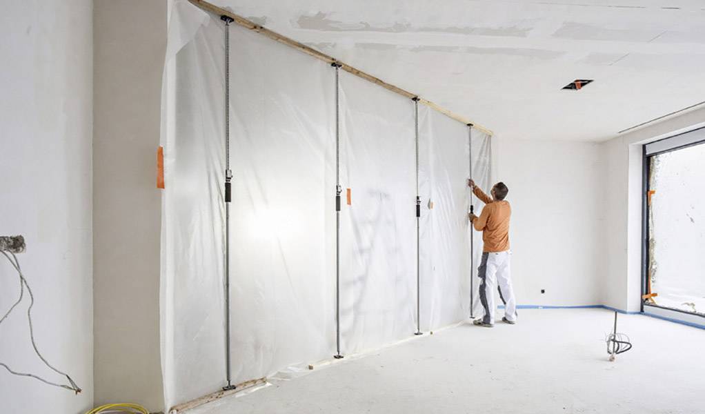 Ein Handwerker befestigt eine Plastikfolie an einer Wand in einem leeren Raum. Die Folie schützt vor Staub und Schmutz.