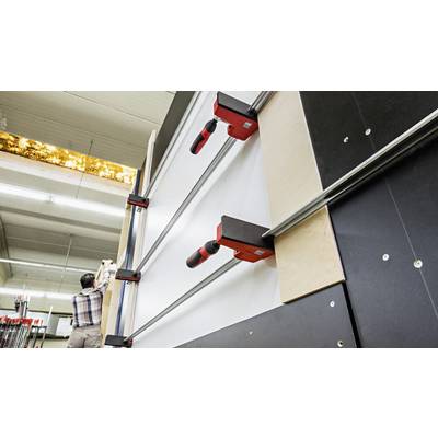 Person klemmt Holzplatte in großer Industriemaschine ein. Einrichtung in einer Werkstattumgebung erkennbar.