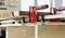 Zwei rote Zwingen halten ein Holzbrett auf einem Tisch fest. Werkzeug dient zur Stabilisierung bei der Holzbearbeitung.