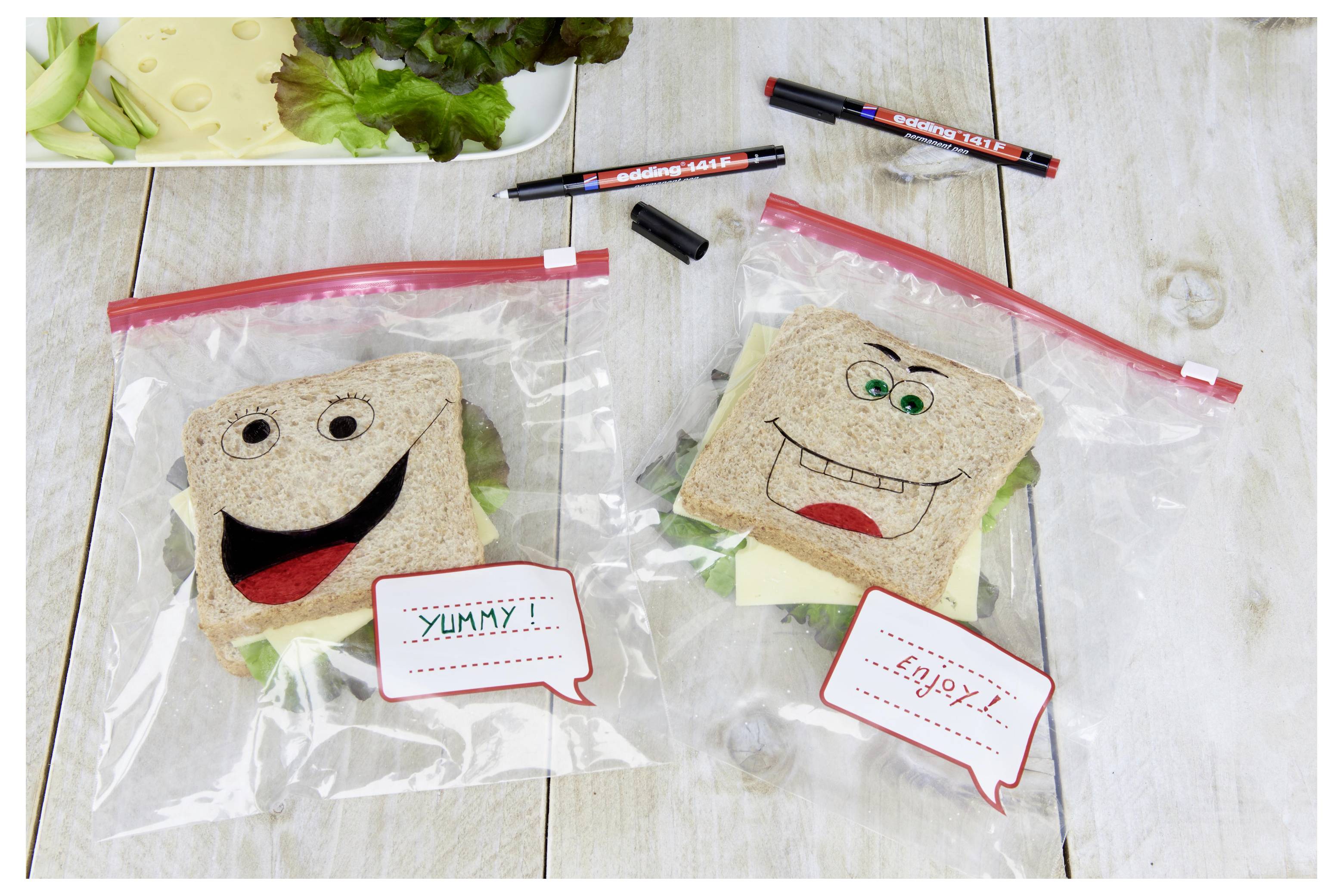 Zwei Hälften von Smiley-Sandwiches in transparenten Reißverschlussbeuteln auf einem Holztisch, jede mit einem beschrifteten Sprechblase; Stifte und ein Tablett mit Salat in der Nähe.