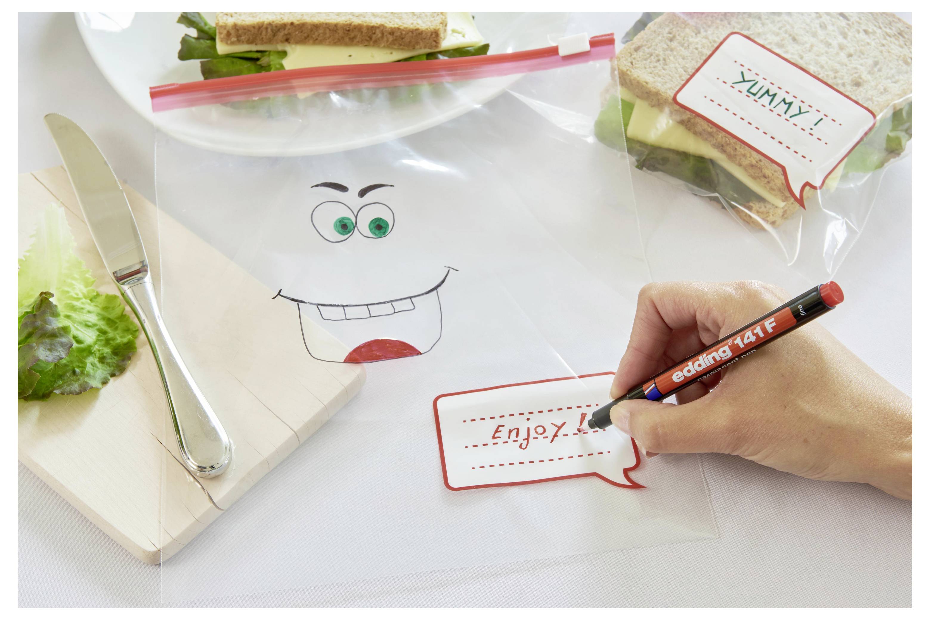 Handschrift "Viel Spaß!" auf einer Plastik-Sandwichtüte mit einem Cartoon-Gesicht, neben einer gepackten Lunchbox und einem Messer auf einem Schneidebrett.