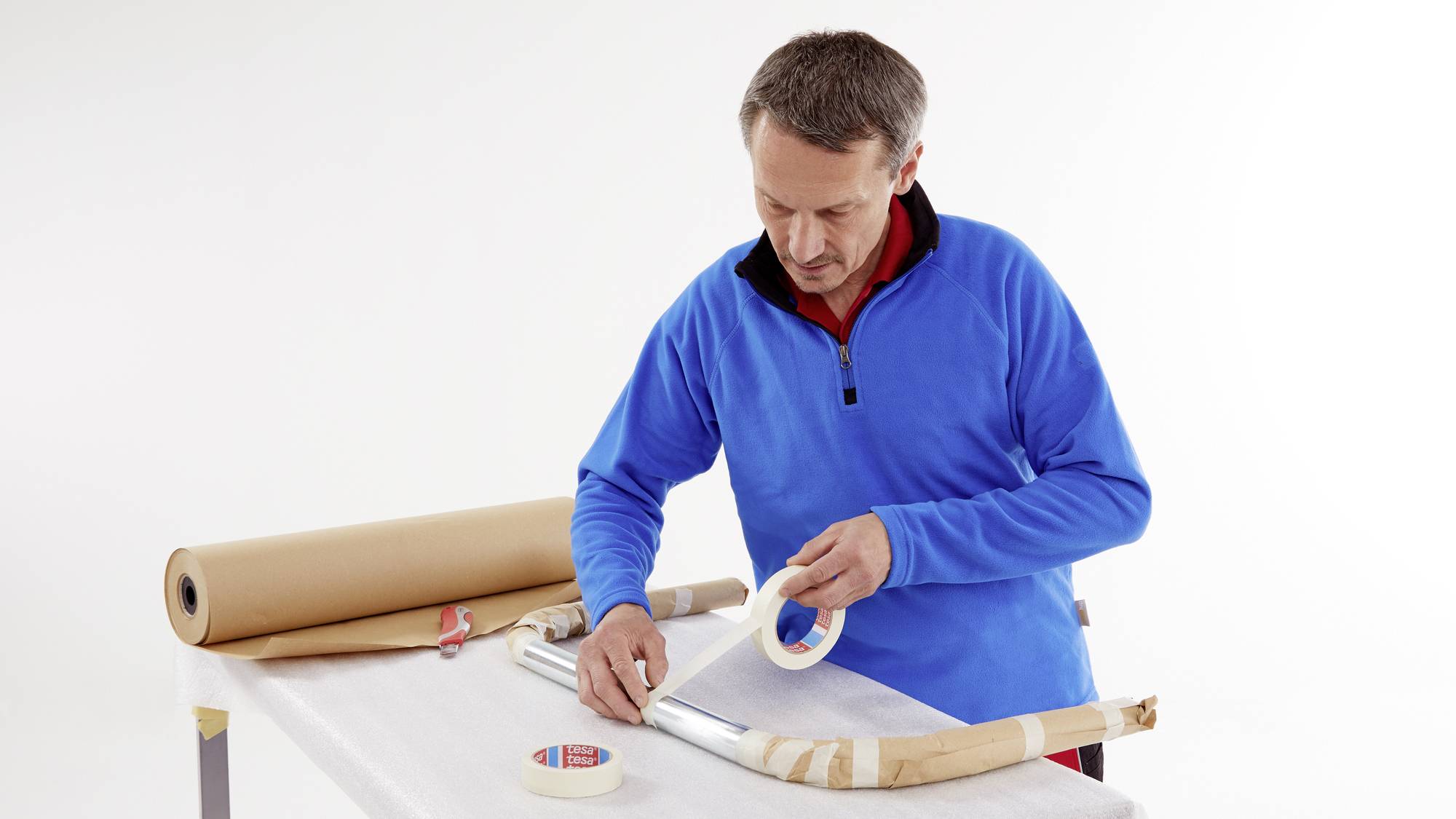 Ein Mann in blauem Pullover verpackt einen Gegenstand mit Papier und Klebeband auf einem Tisch. Rechts von ihm liegt Papierrolle.