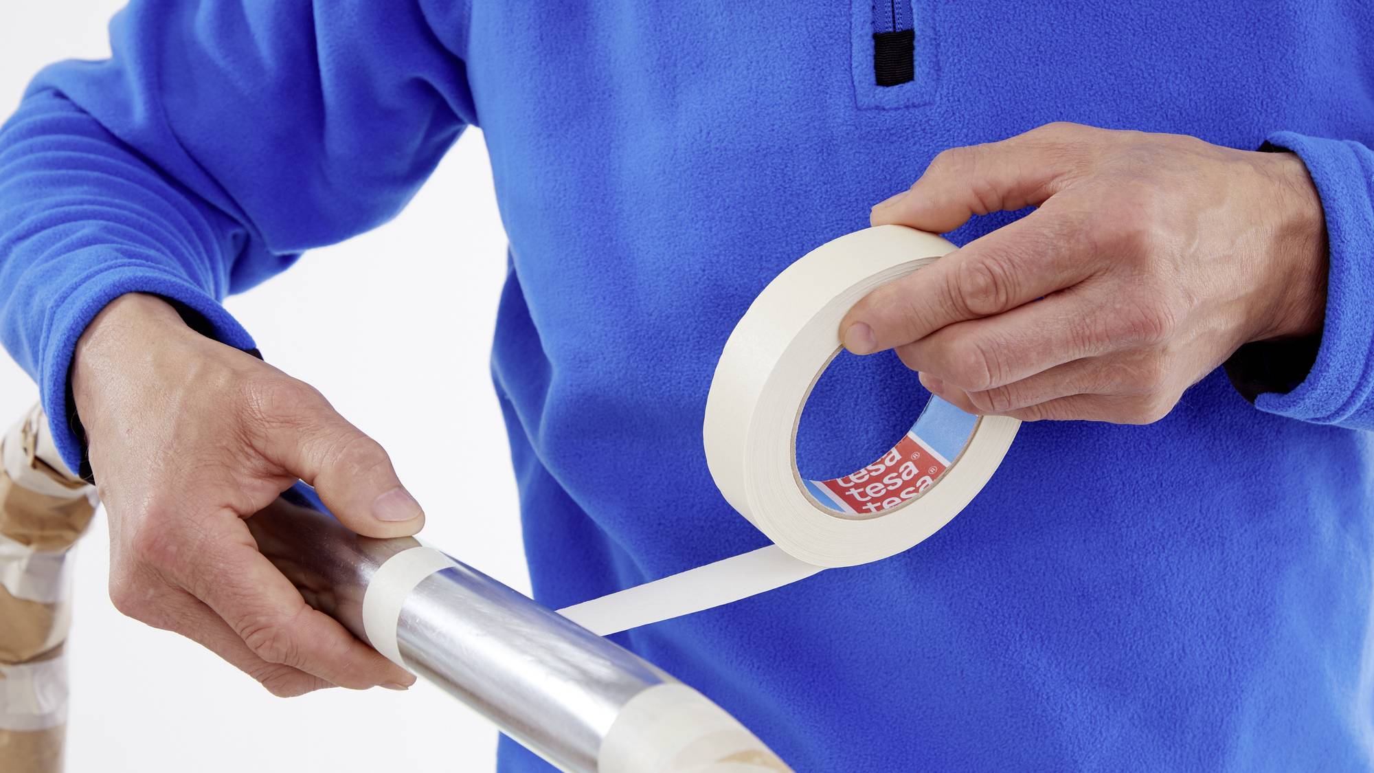 'Person in blauem Pullover umwickelt eine Metallstange mit weißem Klebeband.'