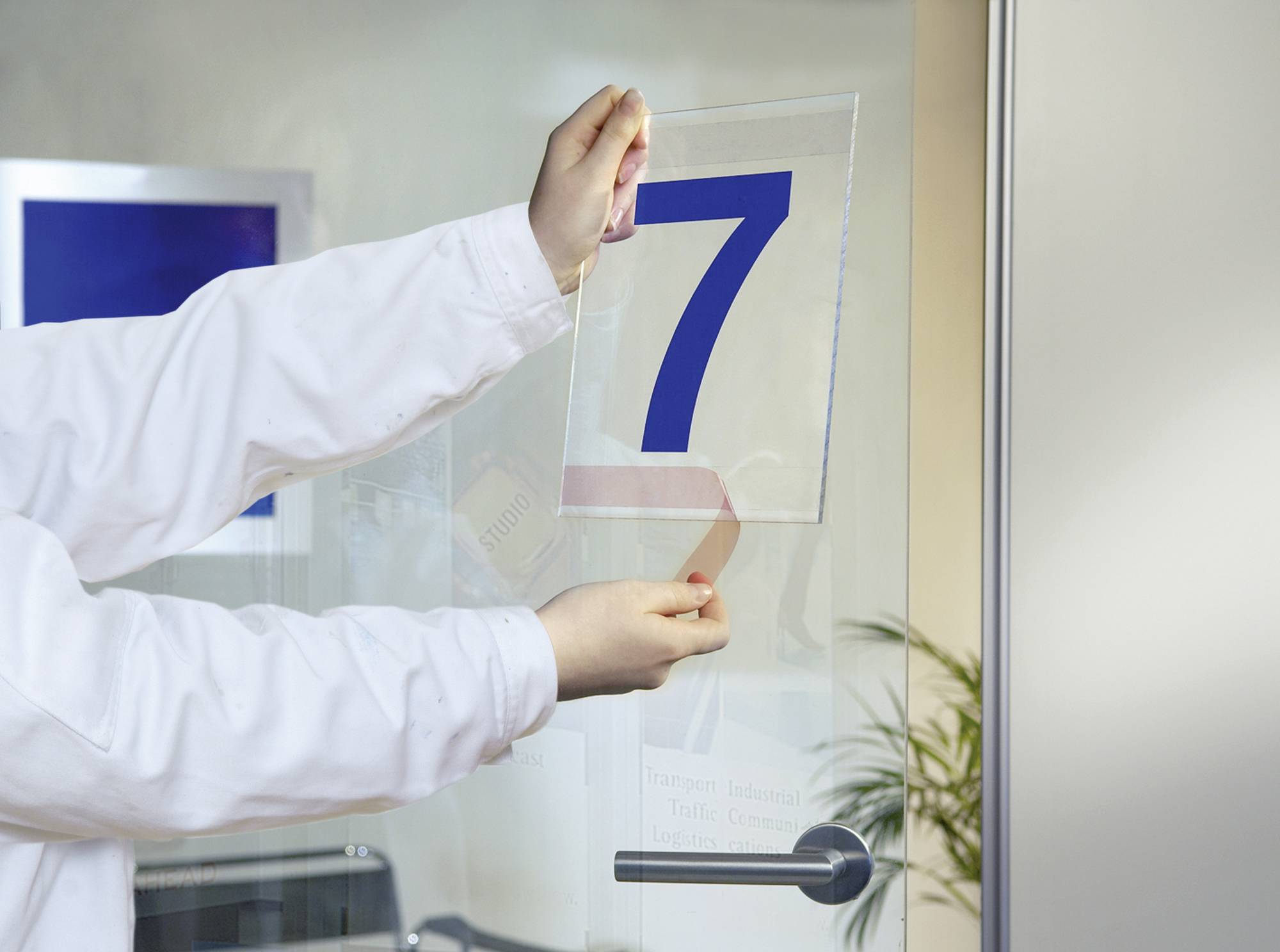 Eine Person in weißem Kittel klebt ein durchsichtiges Schild mit der blauen Nummer 7 an eine Glastür.