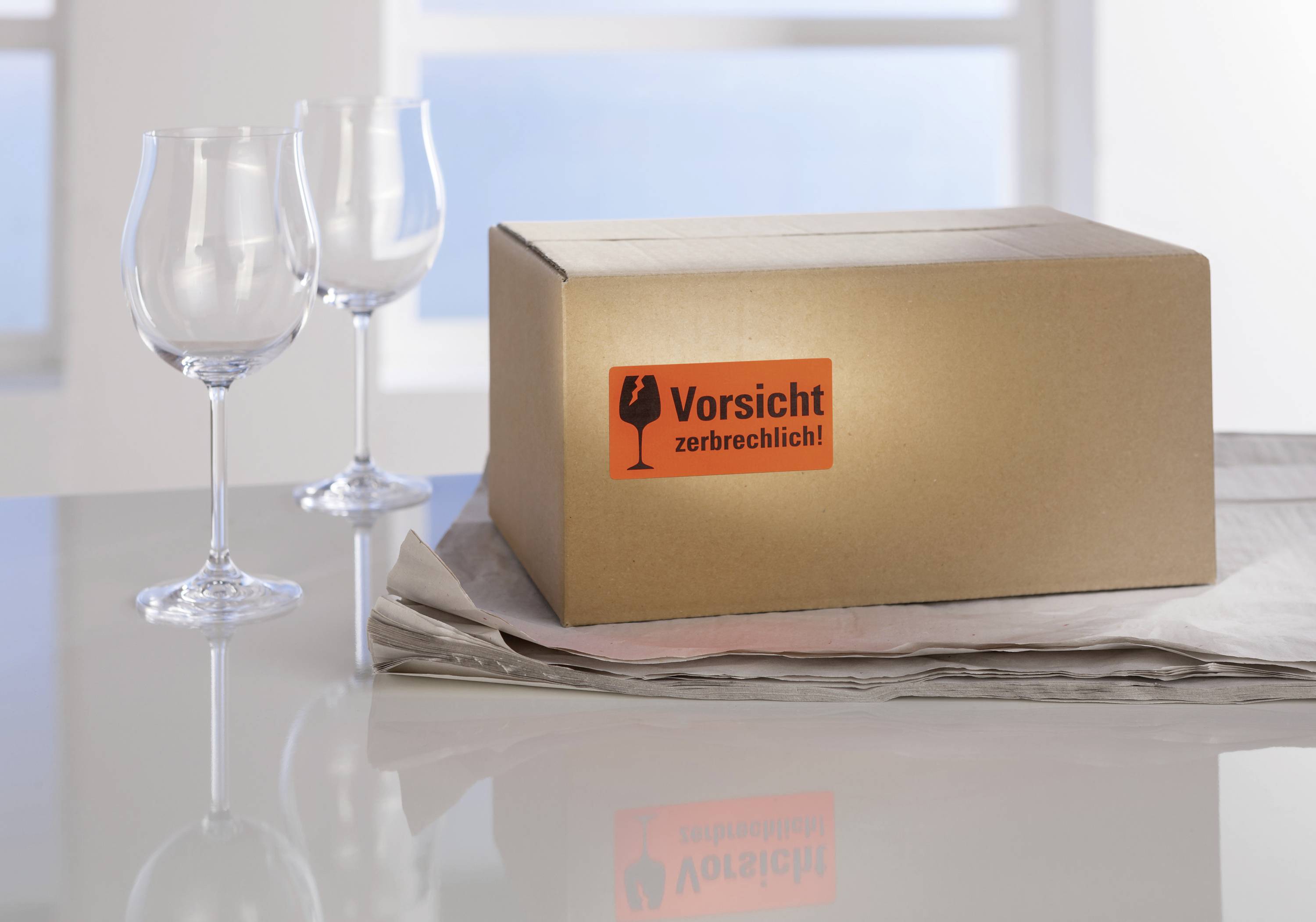 Ein Paket mit Aufkleber 'Vorsicht zerbrechlich!' steht auf einem Tisch mit zwei leeren Weingläsern und Zeitungspapier.