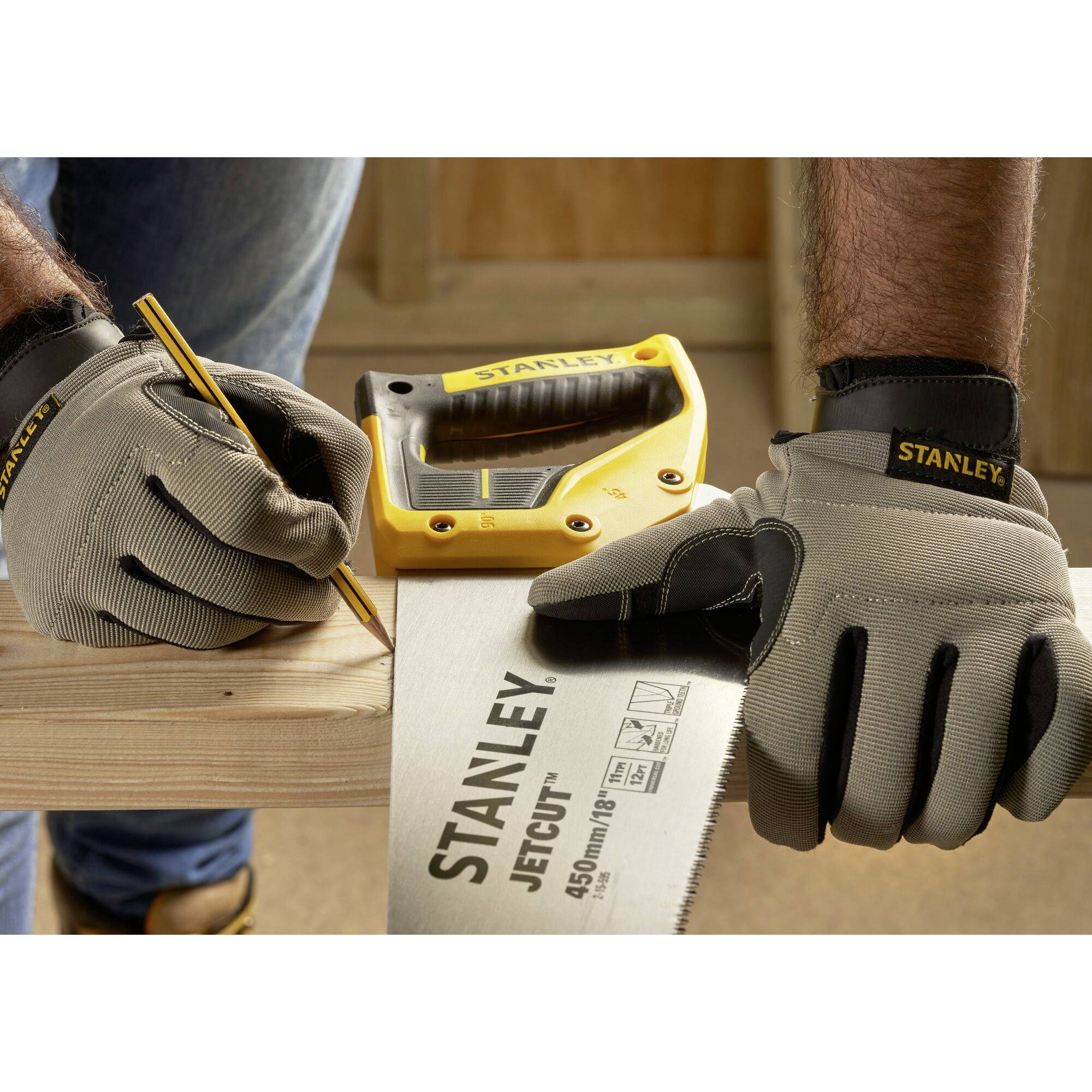 Eine Person mit grauen Handschuhen markiert Holz mit einem Bleistift, während ein Maßband und eine Handsäge auf dem Holz liegen.