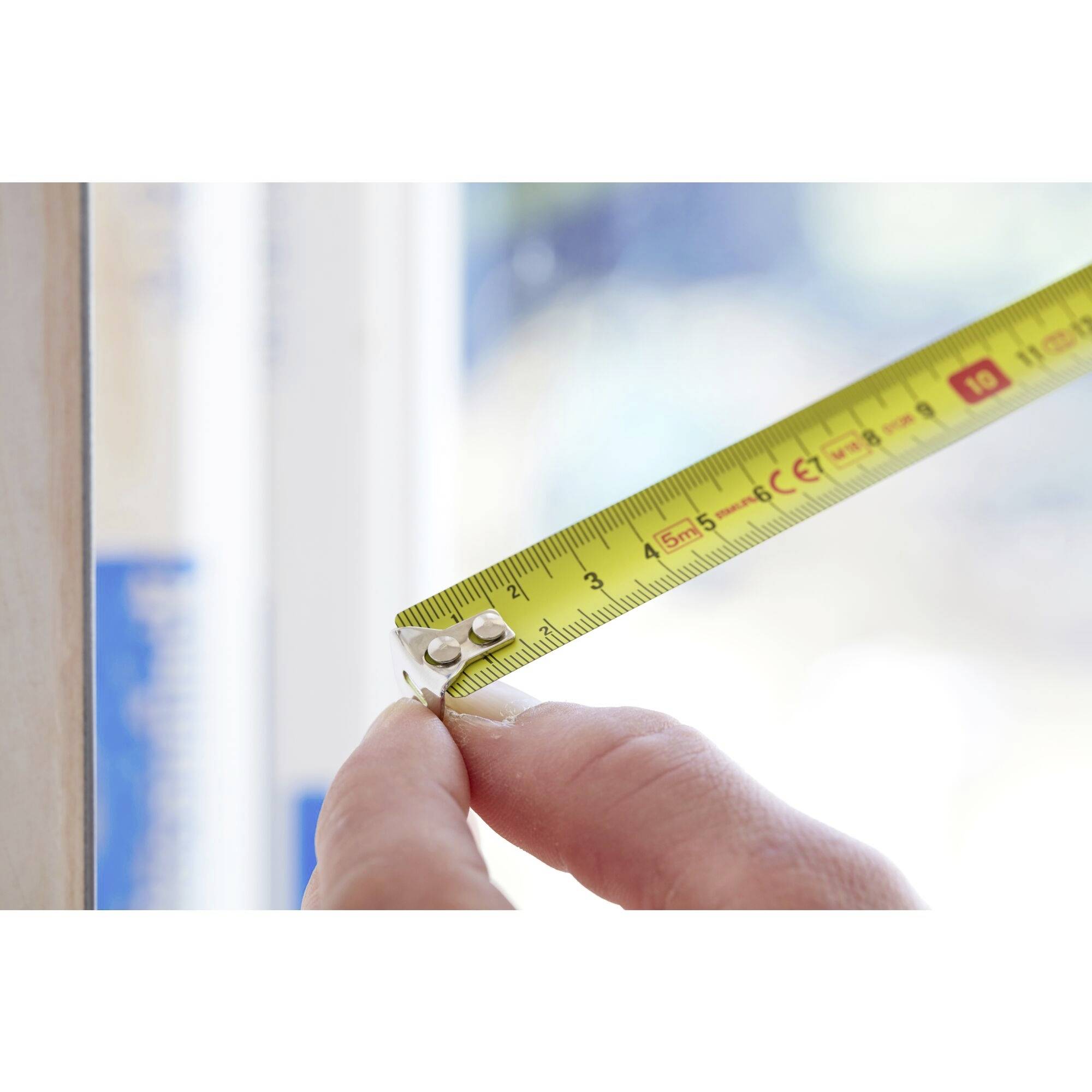Eine Hand hält ein gelbes Maßband, das eine Länge an einem Fensterrahmen misst. Das Maßband zeigt ungefähr 3 Zentimeter an.
