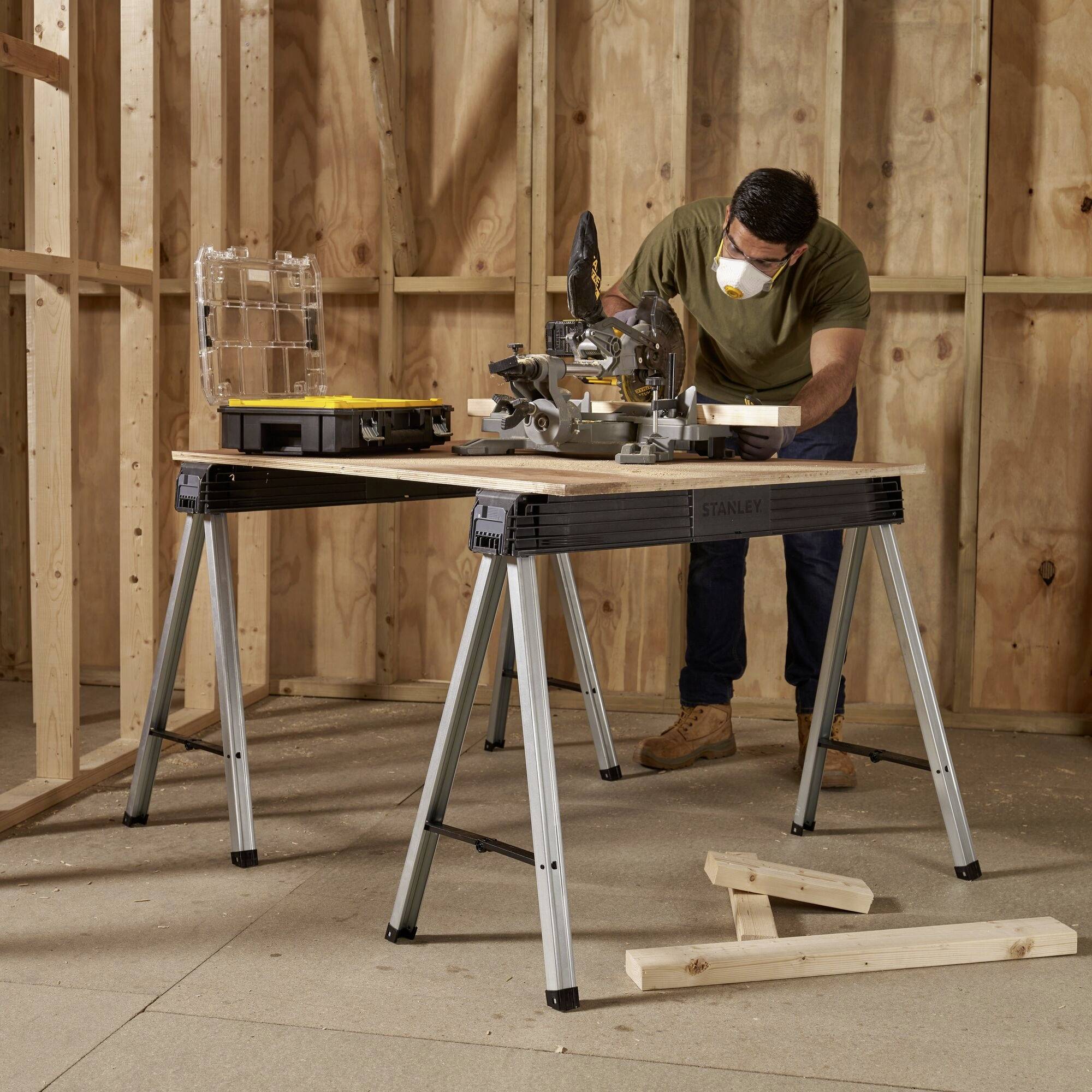 Eine Person arbeitet mit einer Gehrungssäge auf einem Tisch in einer Holzwerkstatt. Sicherheitsausrüstung wird getragen.