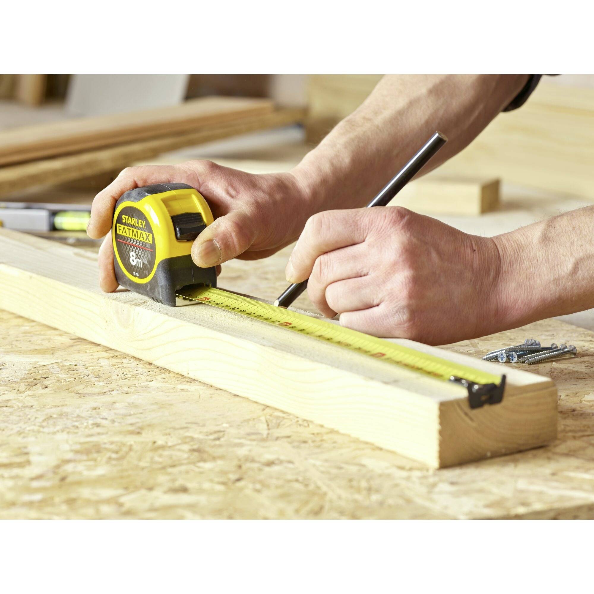 Eine Person misst mit einem Maßband ein Holzbrett und markiert es mit einem Stift auf einem Bautisch. Werkzeuge sind im Hintergrund sichtbar.