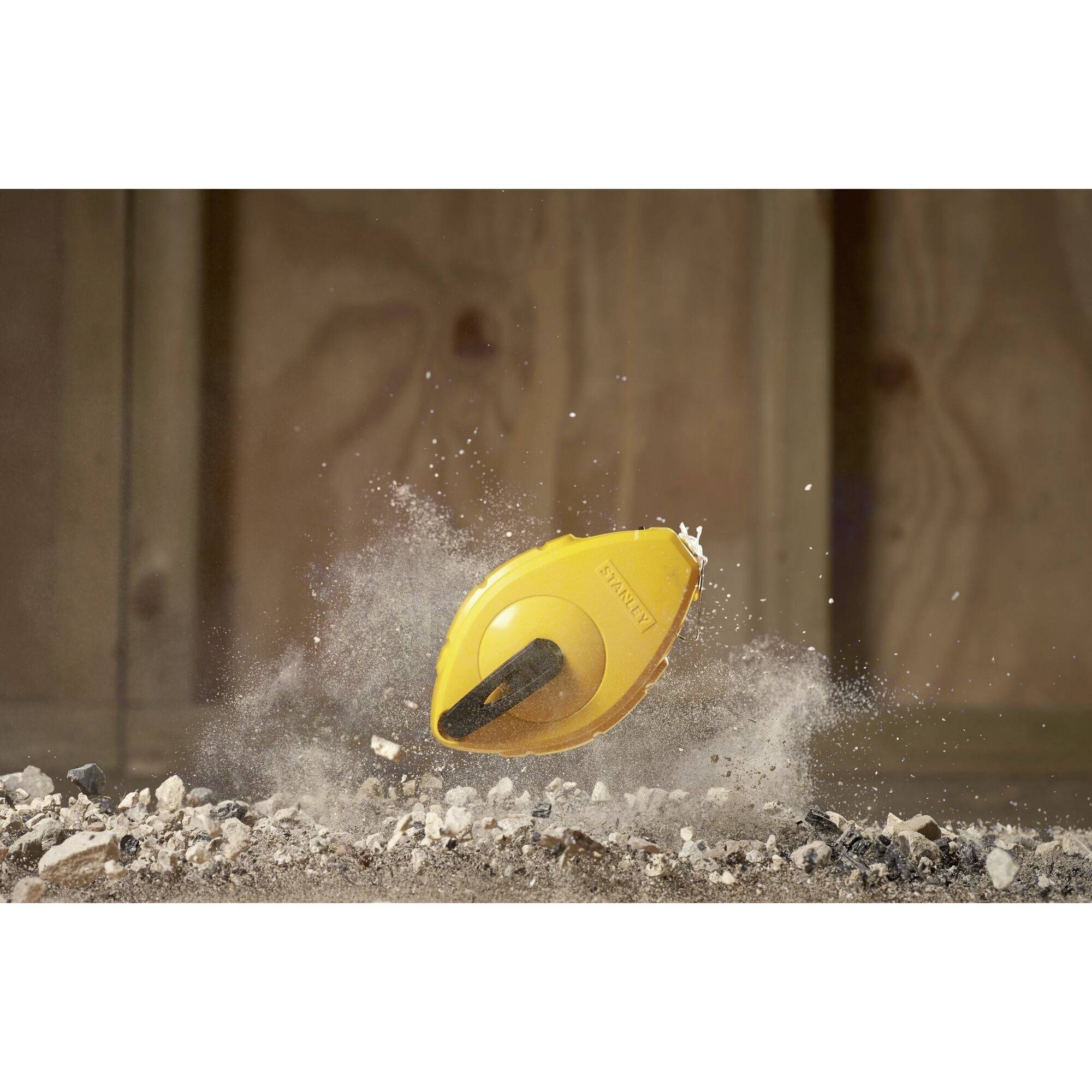 Ein gelbes Maßband fällt auf einen staubigen Boden. Staub und Trümmer fliegen in die Luft. Hintergrund zeigt eine Holzwand.