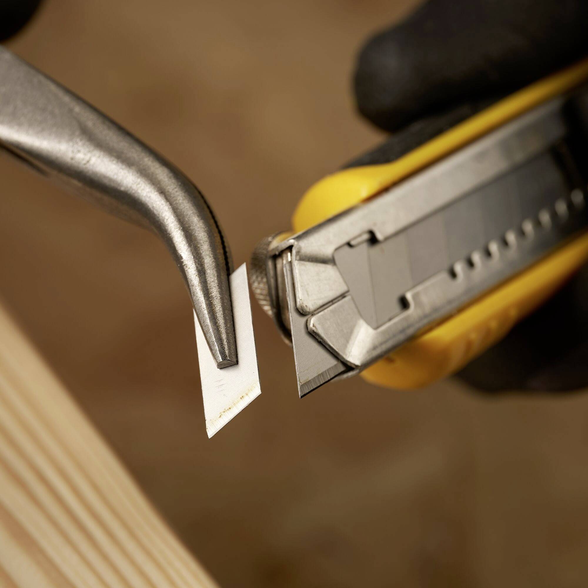 Eine gelbe Cuttermesser-Klinge wird mit einer Zange sicher herausgezogen, vor einem unscharfen Hintergrund aus Holz und Werkstattmaterialien.