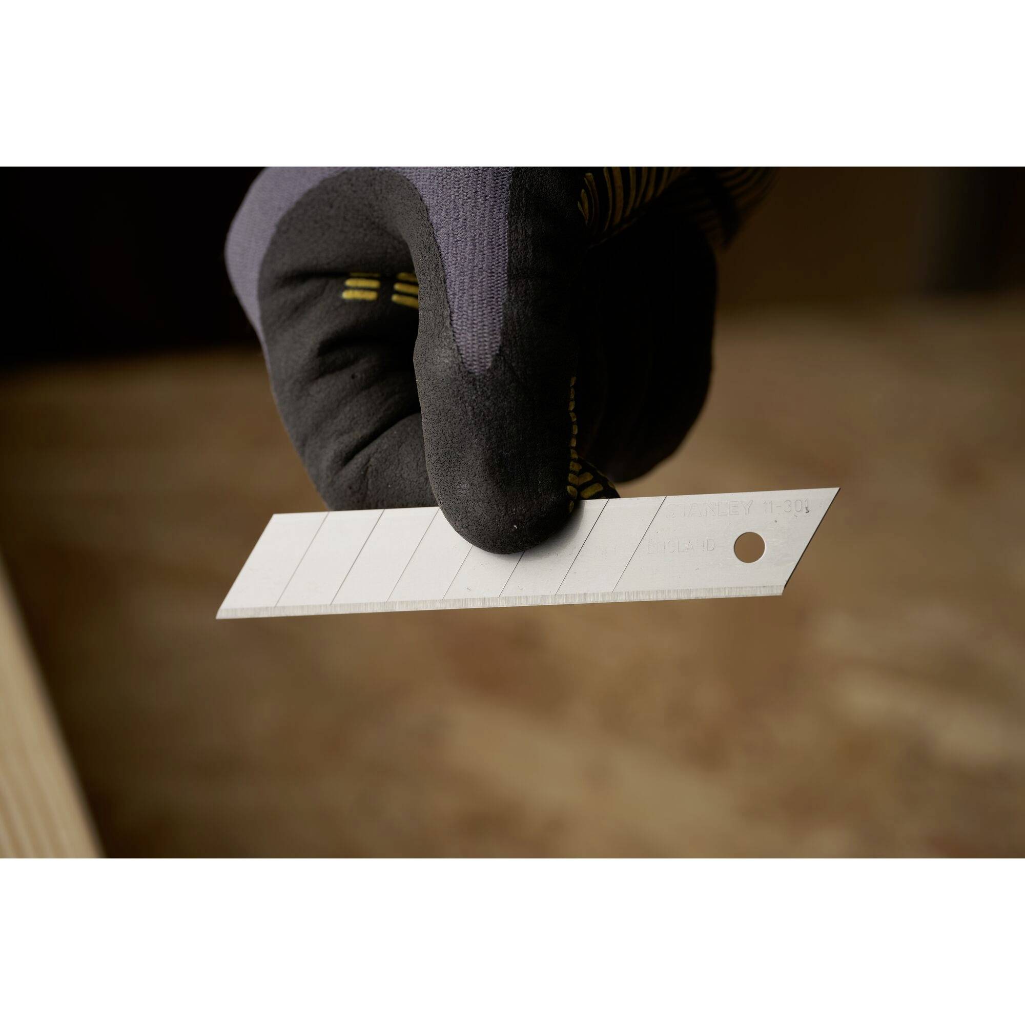 Eine Hand hält eine Ersatzklinge, die zum Schneiden von Materialien verwendet wird. Die Klinge ist silbern und rechteckig mit einer Lochung.