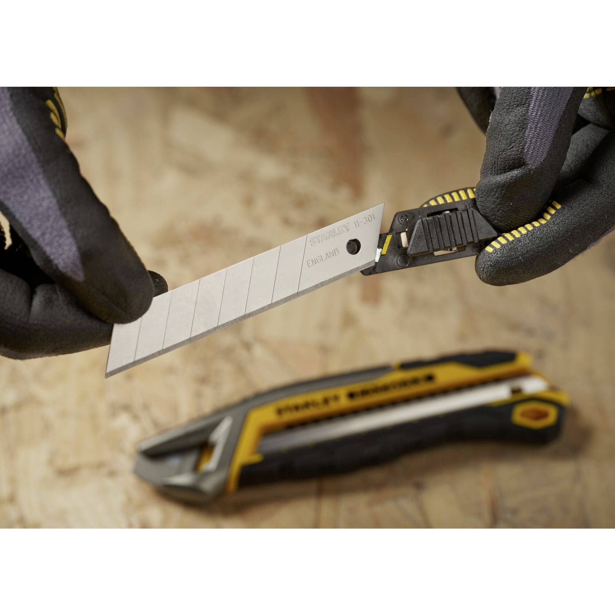 Zwei behandschuhte Hände ziehen eine Klingenhalterung aus einem gelb-schwarzen Cutter-Messer auf einem Holzuntergrund heraus.
