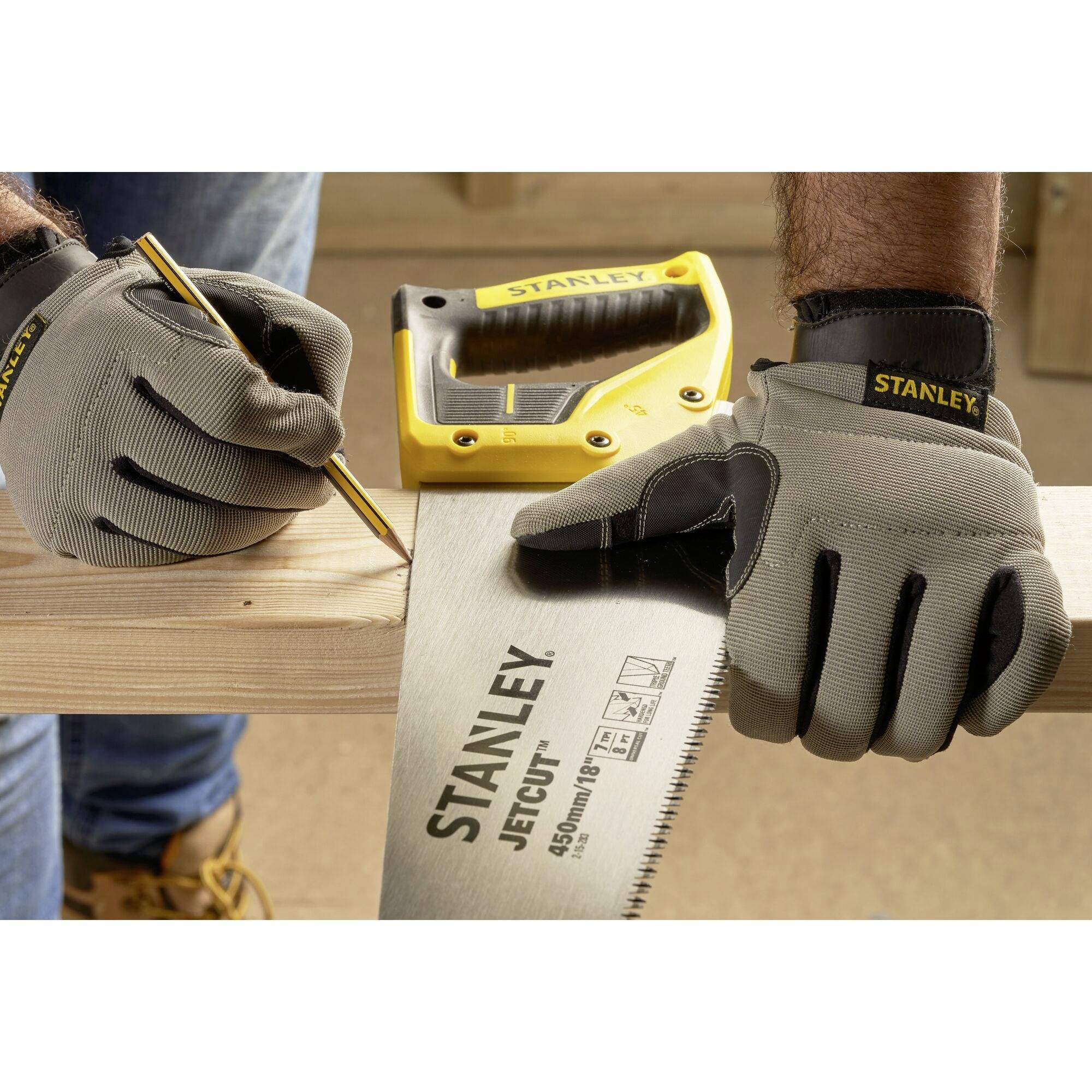 Handwerker mit grauen Handschuhen markiert Holz mit einem Bleistift, hält Säge in der anderen Hand. Stanley-Logo auf der Säge sichtbar.