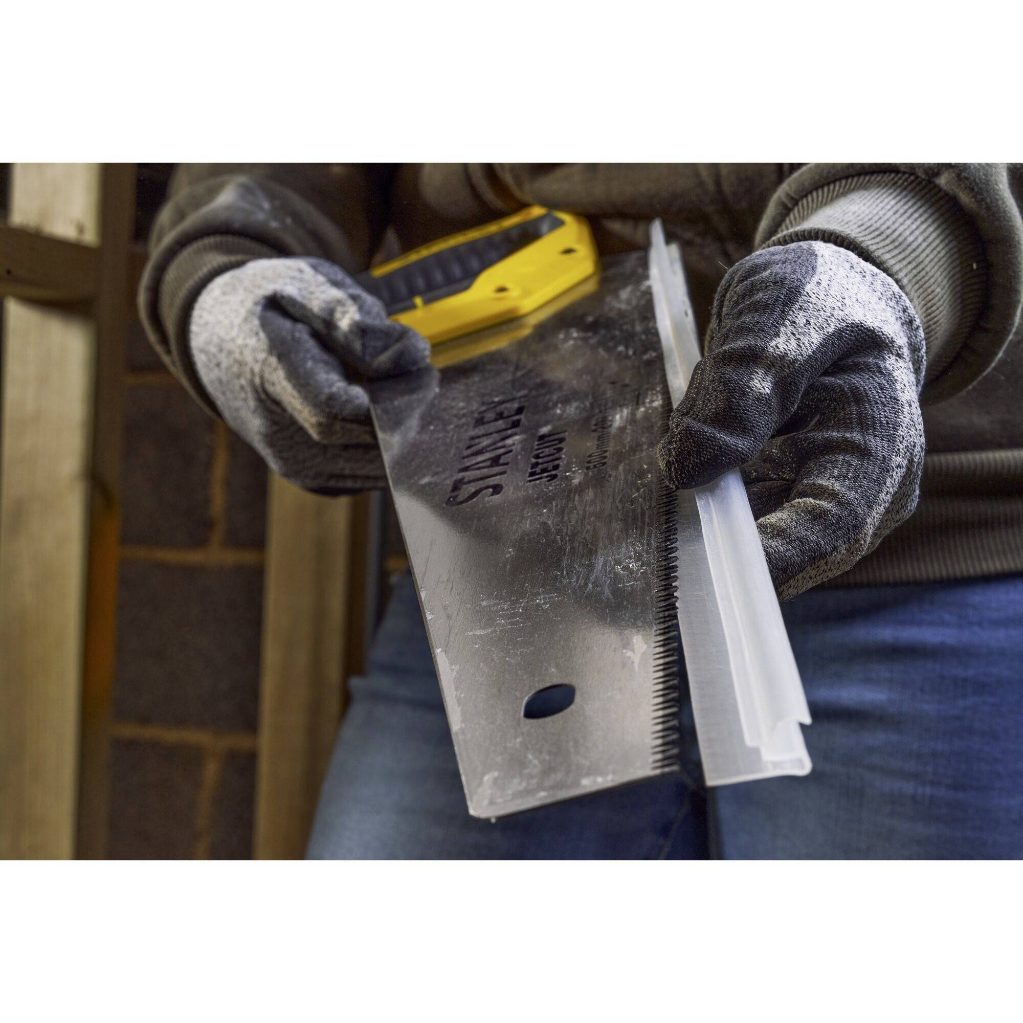 Eine Person in Arbeitshandschuhen hält eine Säge mit Metallblatt, bereit zum Gebrauch. Im Hintergrund sind Ziegel und Holz zu sehen.