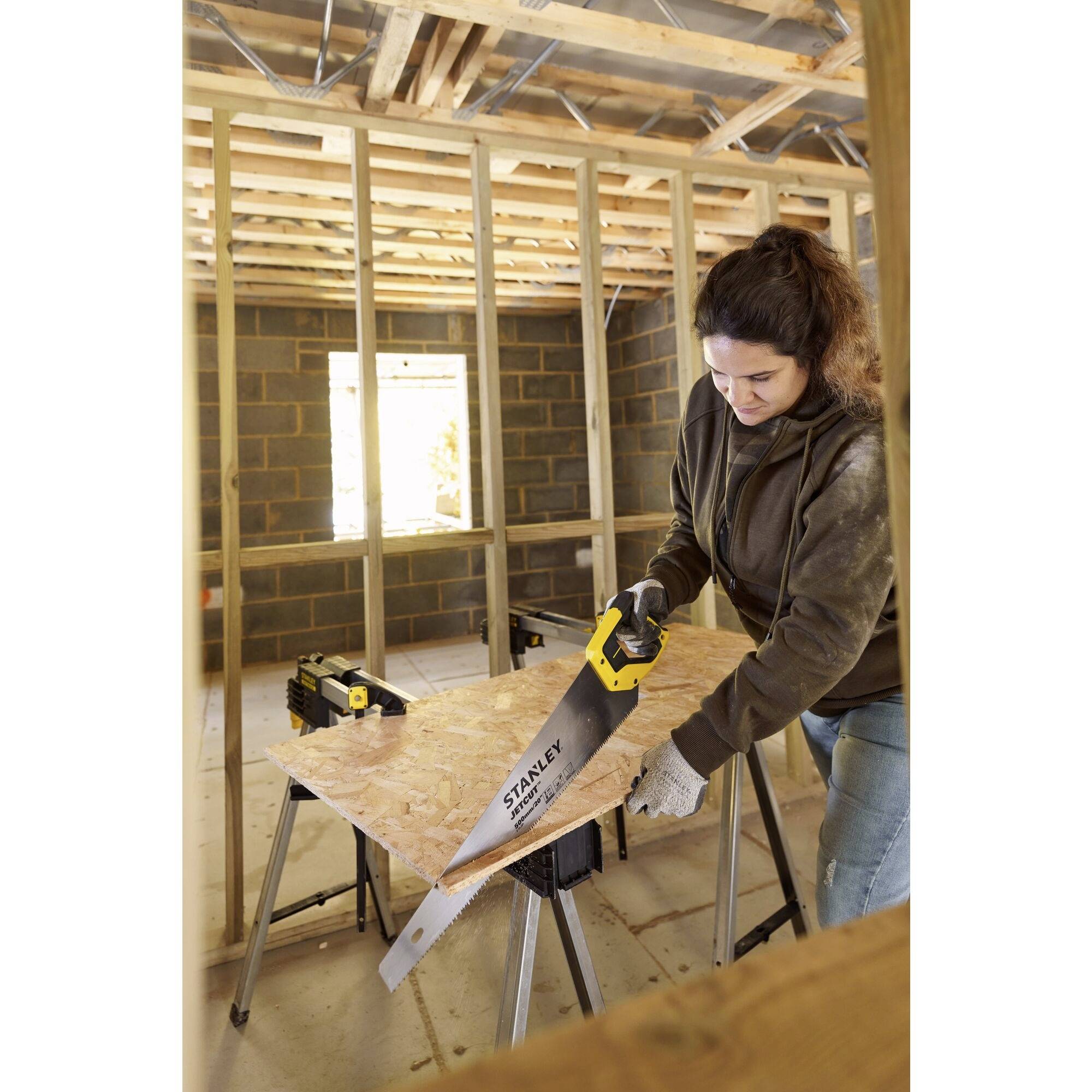 Eine Person sägt mit einer Handsäge ein Holzbrett in einem unfertigen Raum mit sichtbaren Holzstreben und Mauerwerk.
