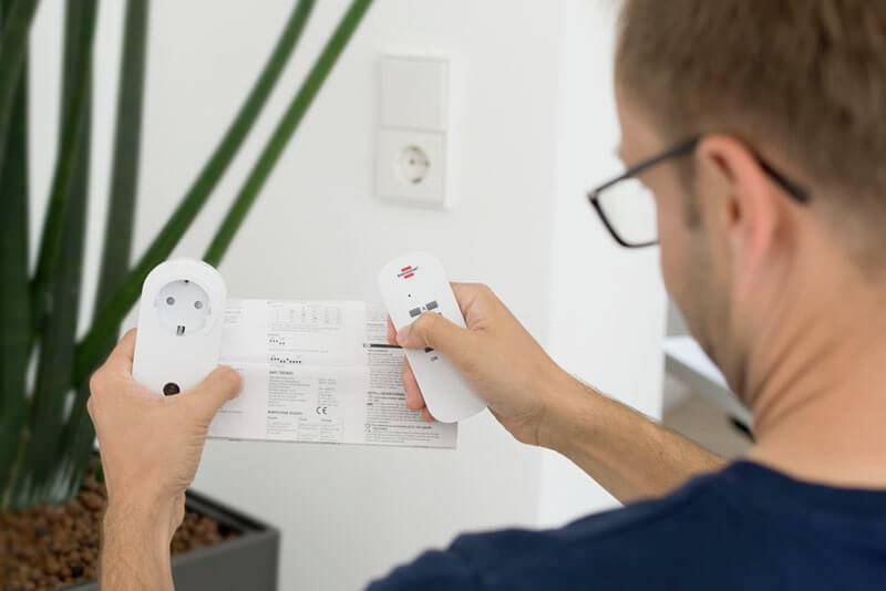 Eine Person hält eine Anleitung und zwei elektronische Geräte, möglicherweise im Zusammenhang mit der Bedienung oder Installation.