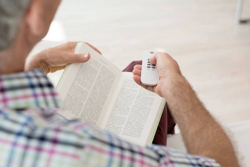 Eine sitzende Person liest ein Buch und hält eine Fernbedienung in der Hand, die auf den Fernseher oder ein Gerät gerichtet ist.