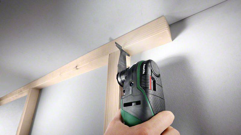 Eine Person nutzt ein elektrisches Multifunktionswerkzeug, um einen Holzrahmen an einer Wand zu bearbeiten.