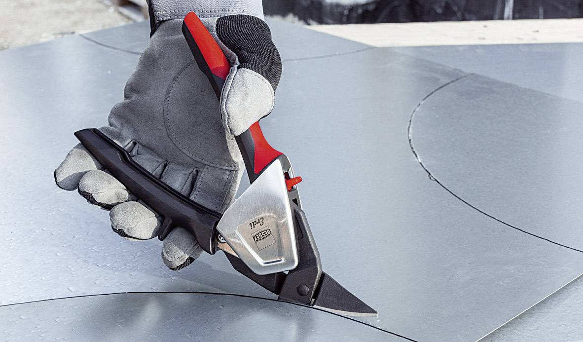 Eine behandschuhte Hand benutzt eine Blechschere, um eine Markierung auf einem Metallblech zu schneiden.