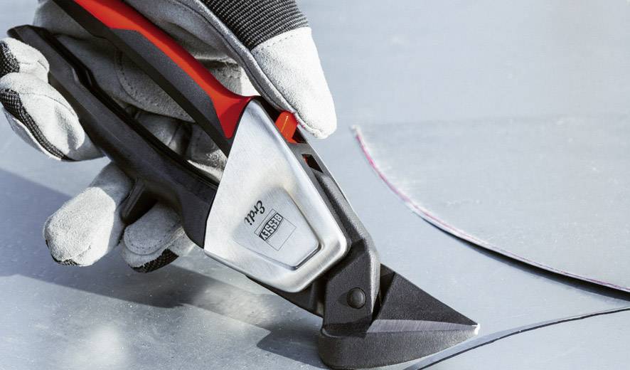 Eine Hand mit Handschuhen schneidet mit einer Metallschere präzise durch ein dünnes, gebogenes Metallblech.