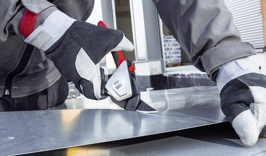 Handwerker in grauer Arbeitskleidung schneidet mit einem Messer ein Metallblech. Arbeitsumgebung im Hintergrund.