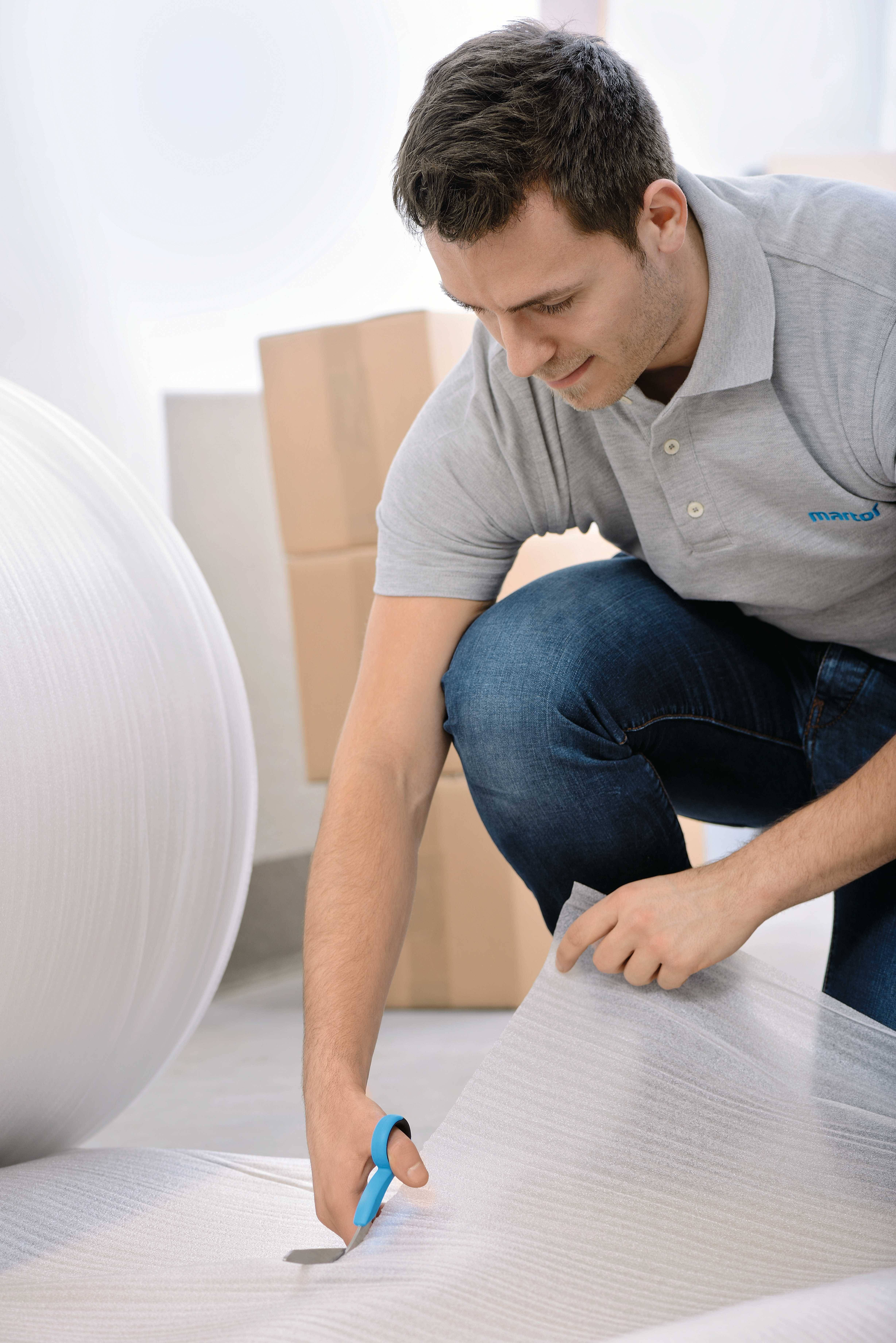 Mann in Jeans und Polohemd schneidet mit einer Schere Verpackungsmaterial. Im Hintergrund sind Kartons zu sehen.