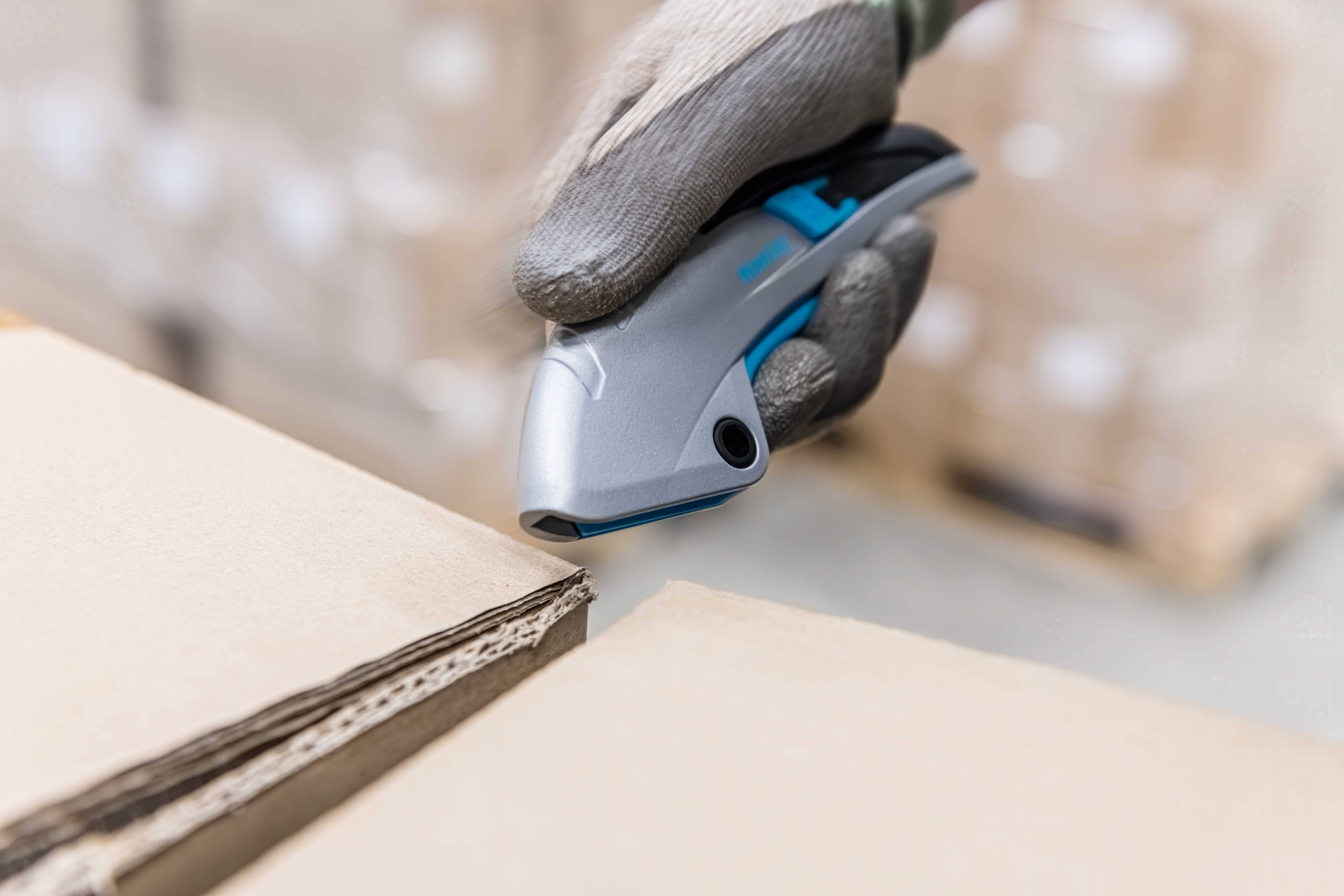 'Hand in Handschuhen nutzt ein Verpackungsmesser, um einen Karton zu öffnen. Kartons im Hintergrund deuten auf Lagerumgebung hin.'