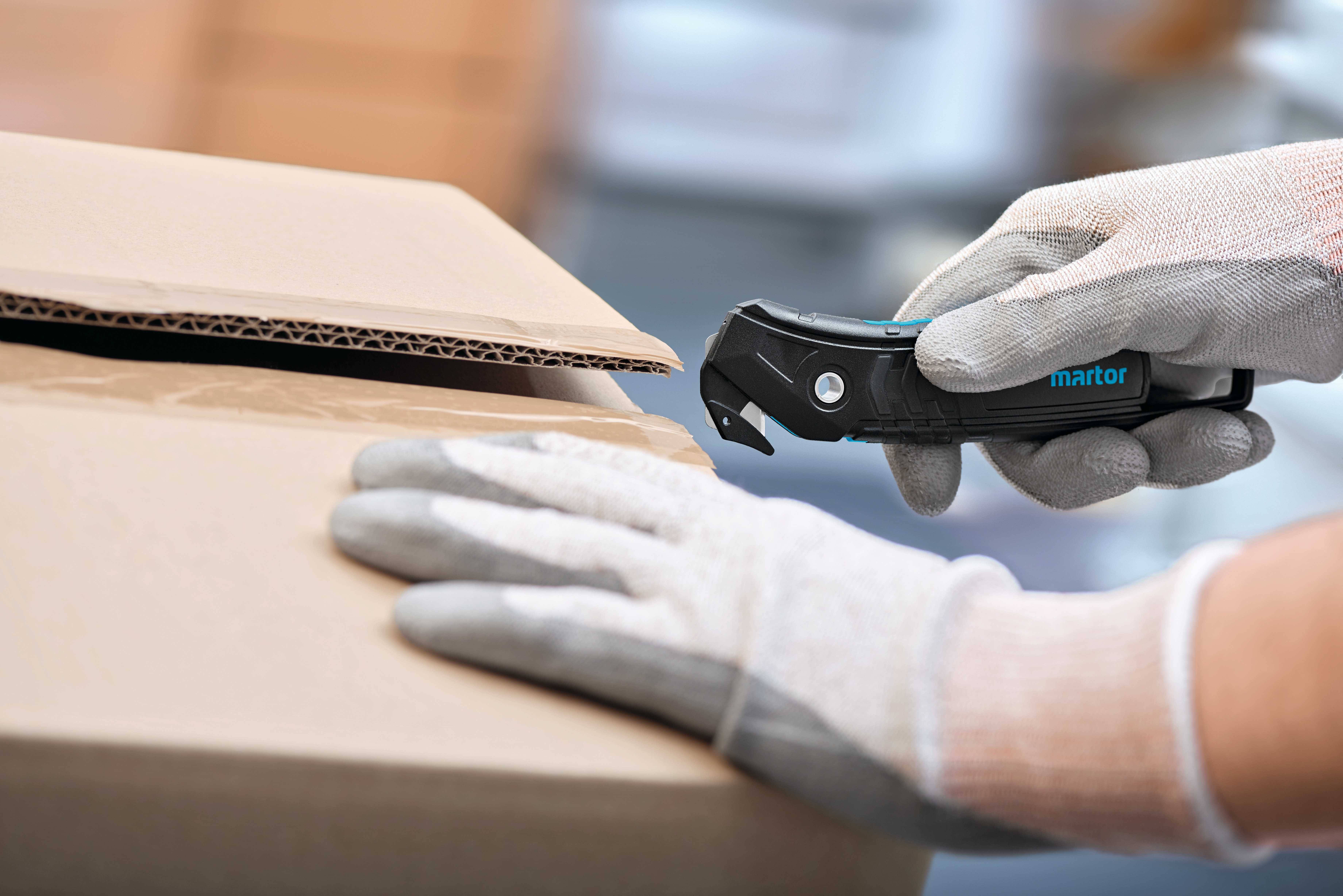 Eine Person öffnet mit einem Sicherheitsmesser einen Karton. Die Hände sind mit Arbeitshandschuhen geschützt.