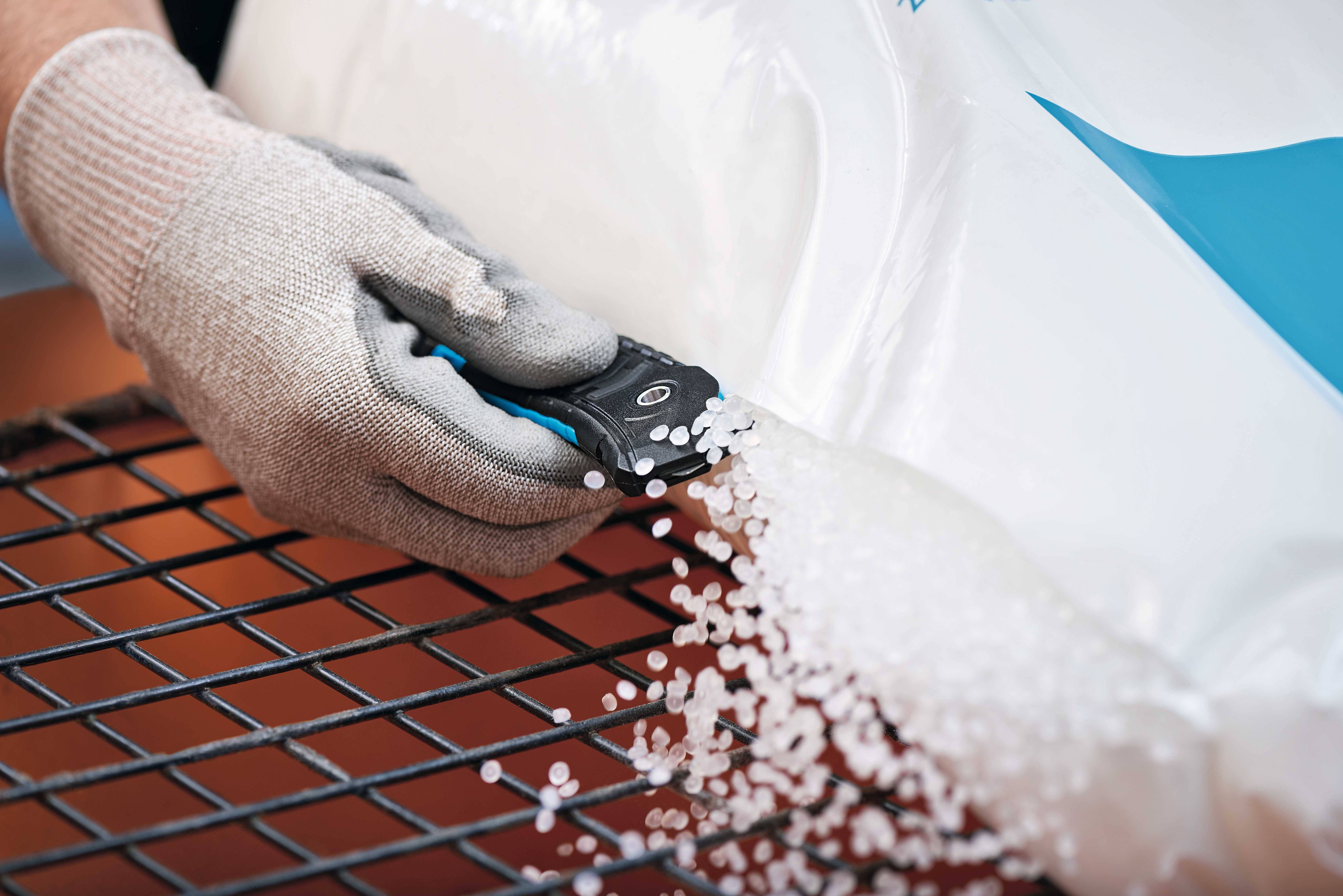 Eine behandschuhte Hand schneidet einen Plastikbeutel auf, aus dem weiße Kunststoffpellets auf ein Gitter rieseln.