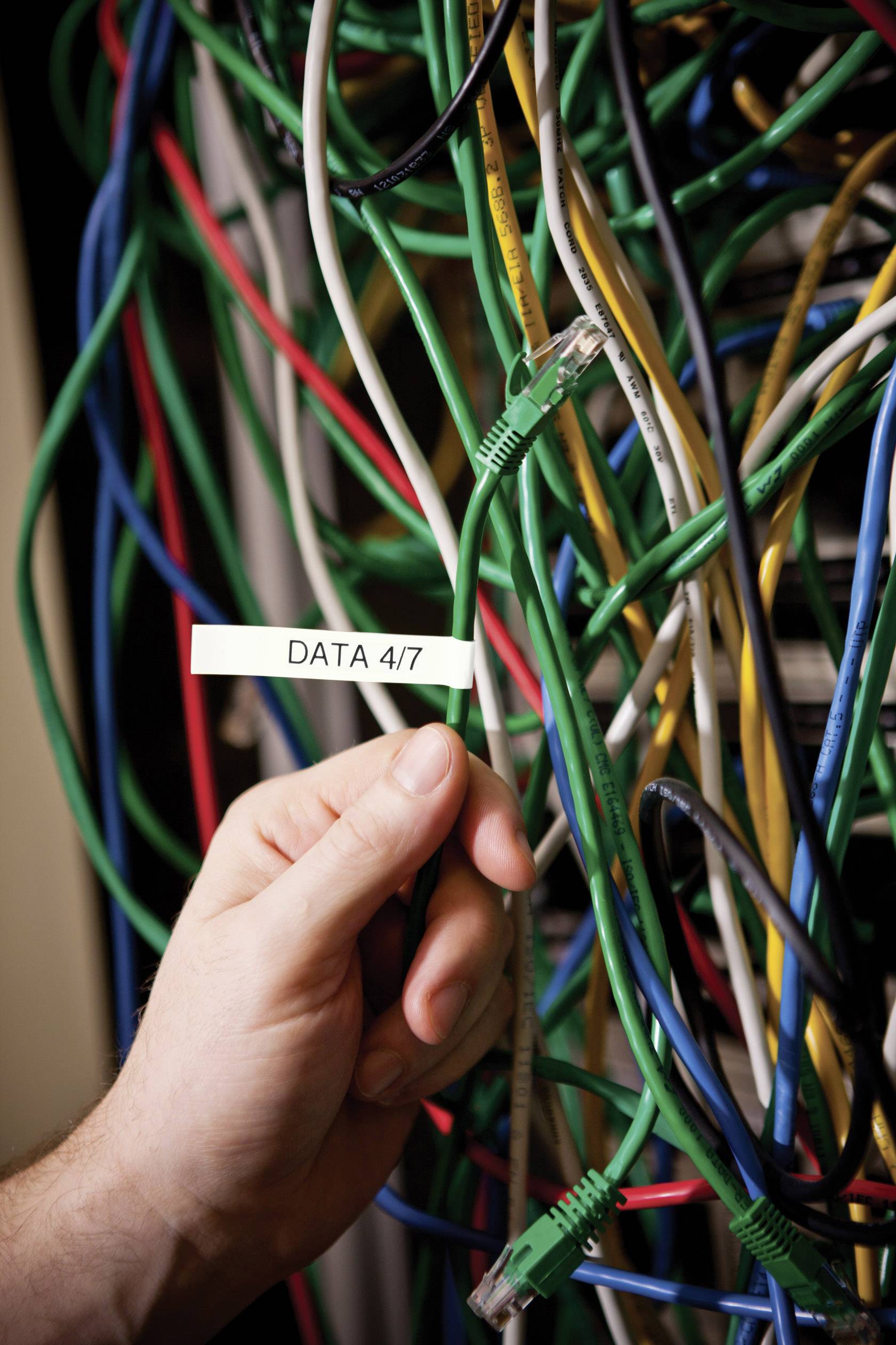 Eine Hand hält ein Kabel, beschriftet mit 'DATA 4/7', inmitten eines Bündels verschiedenfarbiger Netzwerkdrähte.