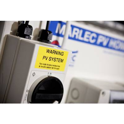 Warnschild neben PV-System-Komponente mit Warnung 'WARNING PV SYSTEM'. Im Hintergrund ein unscharfer Marlec PV-Aufkleber. Hauptfokus auf Sicherheitshinweis.