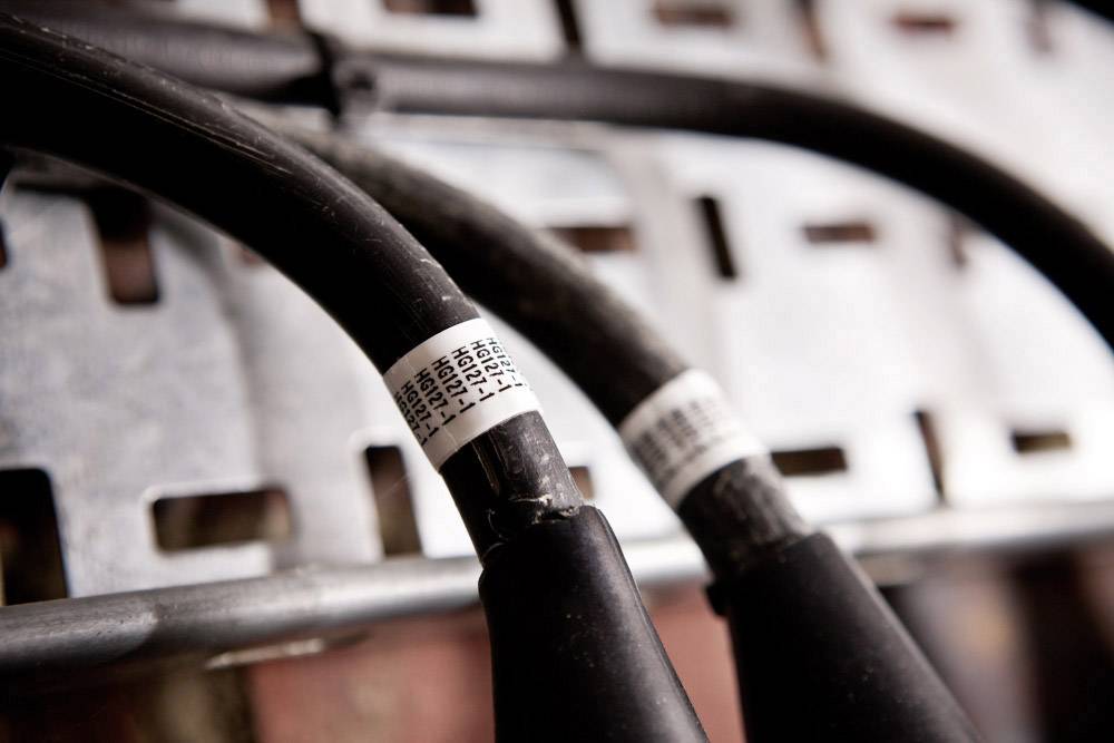 Zwei dicke schwarze Kabel mit weißen Etiketten sind nebeneinander auf einer Metallhalterung montiert. Sie befinden sich in einem industriellen Umfeld.