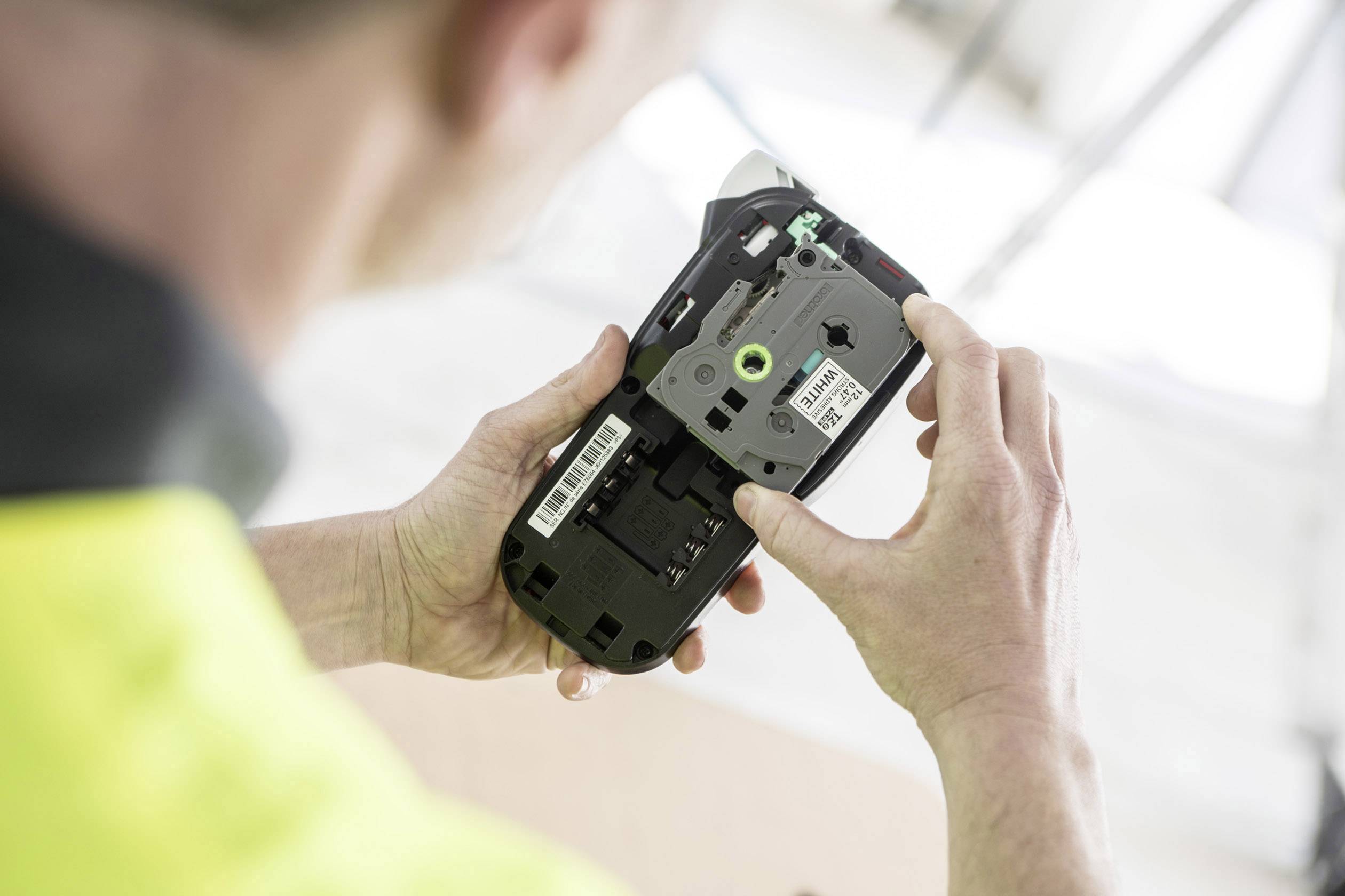 Eine Person hält ein geöffnetes elektronisches Gerät, vermutlich zur Inspektion oder Reparatur, erkennbar sind Drähte und Barcode-Etiketten.