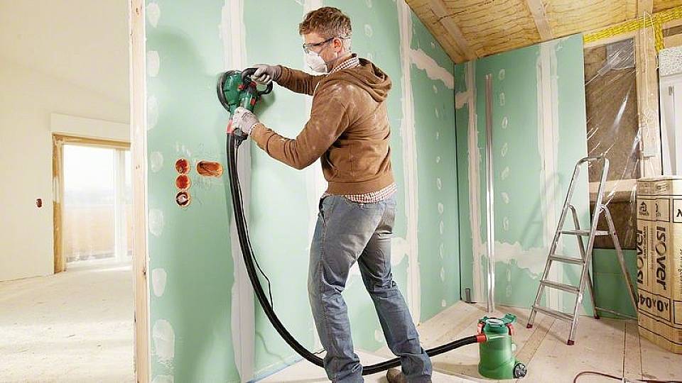 Eine Person schleift eine grüne Gipskartonwand mit einem elektrischen Schleifgerät in einem Raum, der renoviert wird.
