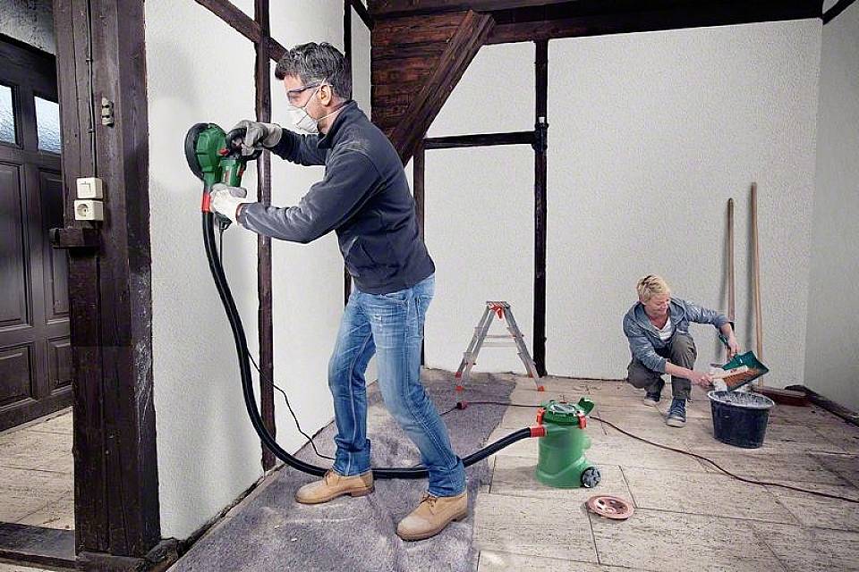 Ein Mann benutzt ein Schleifgerät an einer Wand, während eine weitere Person im Hintergrund Werkzeug vorbereitet. Renovierungsszene in einem Raum.