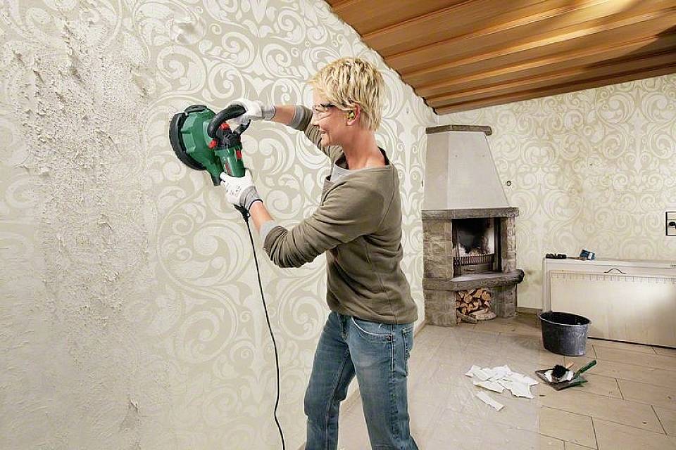Eine Person schleift eine Wand mit einem Elektrowerkzeug in einem Wohnzimmer. Neben der Person steht ein Eimer mit Werkzeugen.