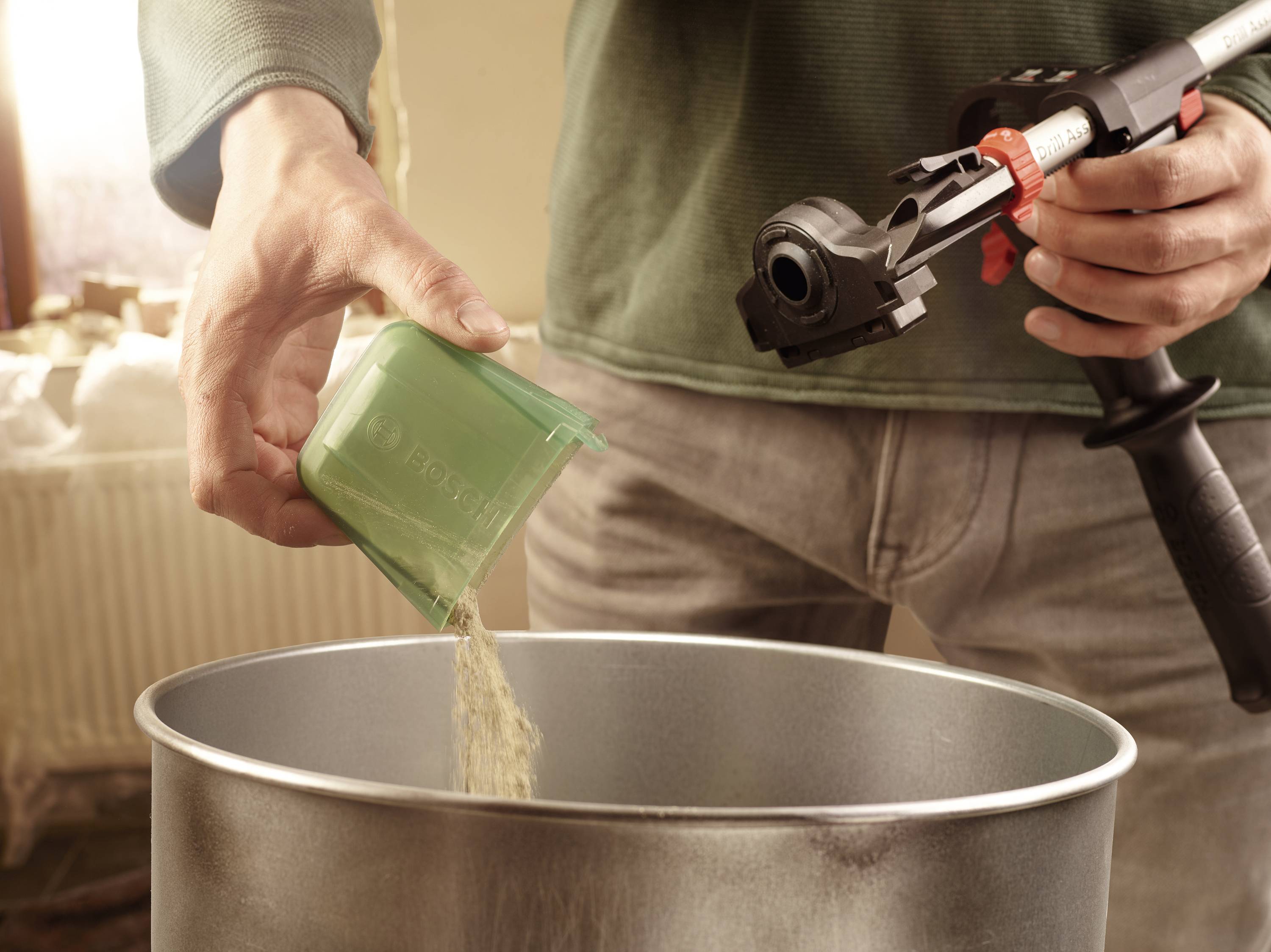 Eine Person füllt einen grünen Becher mit Pulver in einen großen Metalleimer. Daneben wird ein Gerät mit einem Griff gehalten.