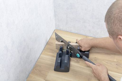 Ein Mann schneidet mit einer Handsäge ein Holzstück auf einem Tisch. Er benutzt eine Gehrungslade, um einen genauen Winkel zu erzielen.