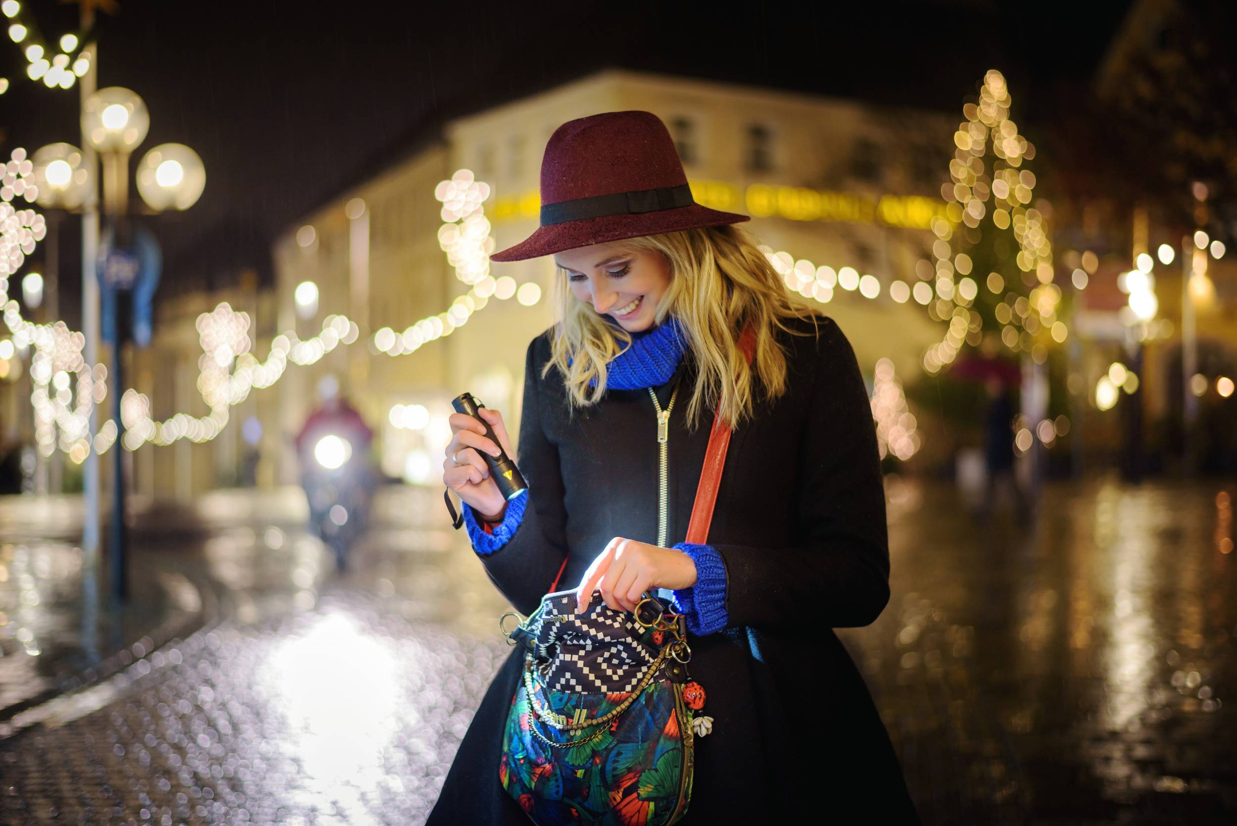 Eine Frau mit Hut leuchtet mit einer Taschenlampe in ihre Handtasche auf einer beleuchteten, nächtlichen Stadtstraße.