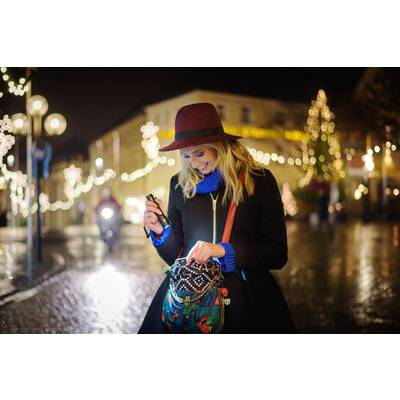 Eine Frau mit Hut leuchtet mit einer Taschenlampe in ihre Handtasche auf einer beleuchteten, nächtlichen Stadtstraße.