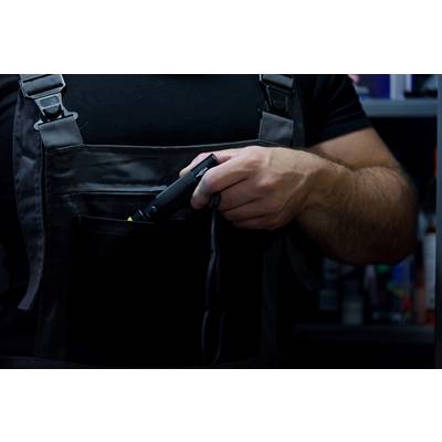 Eine Person steckt ein Werkzeug in die Tasche einer Arbeitslatzhose. Sie befindet sich vor einem unscharfen Hintergrund mit Regalen.