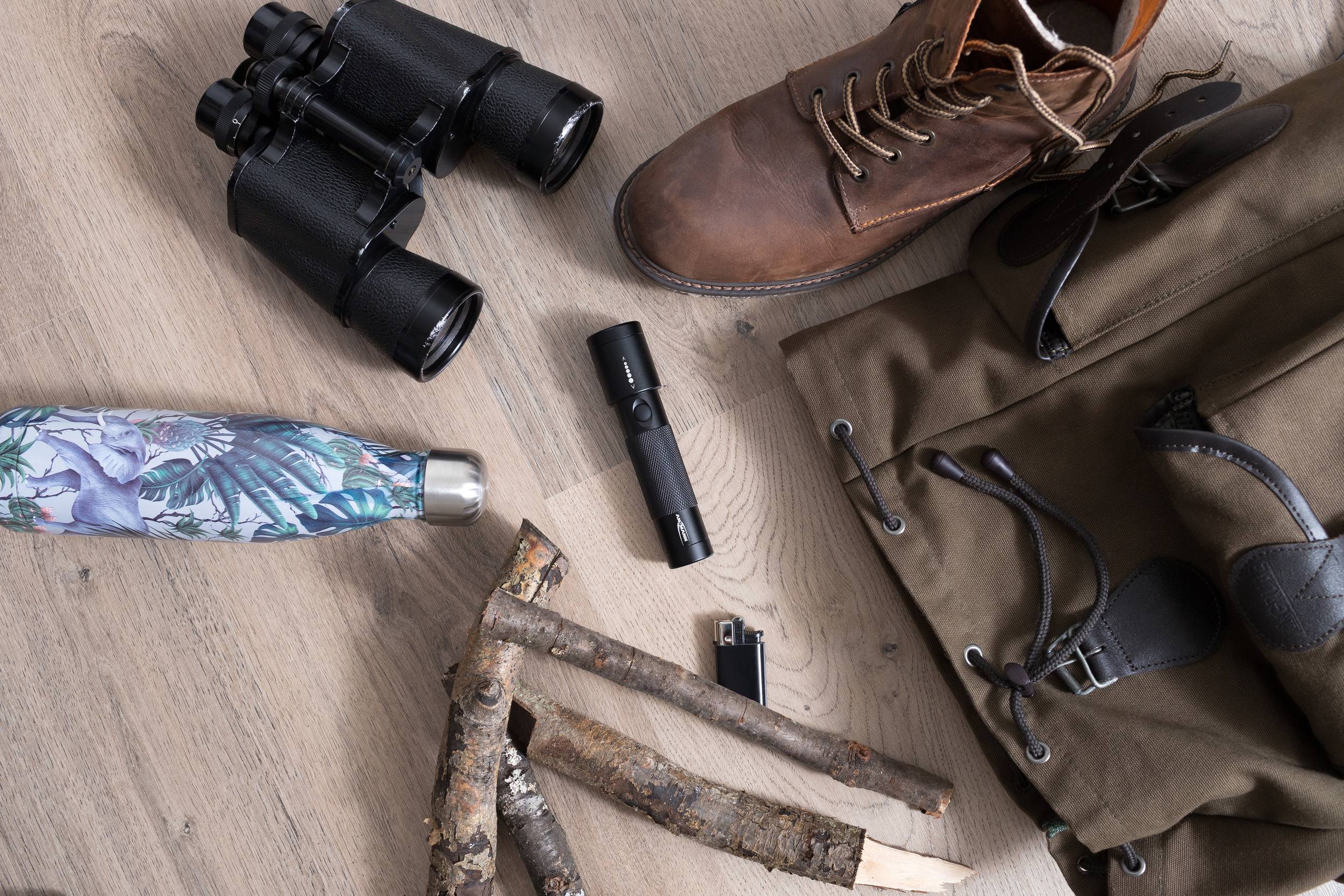 Wanderausrüstung auf Holzboden: Fernglas, Wanderstiefel, Feldflasche, Taschenlampe, Stöcke, Feuerzeug, olivfarbene Tasche.