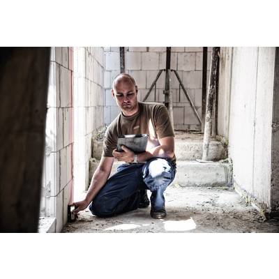Ein Mann kniet in einem Rohbau und misst mit einem Gerät die Wände. Er hält einen Klemmbrett und trägt Arbeitskleidung.