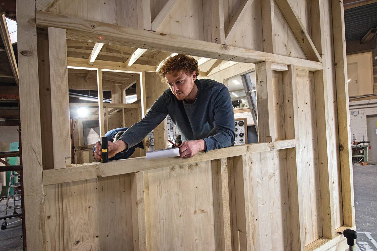 Eine Person arbeitet an einem Holzrahmen in einer Werkstatt, verwendet ein Maßband und macht Markierungen auf dem Holz für einen präzisen Schnitt.