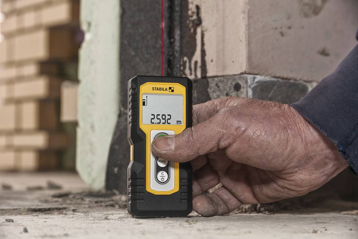 Eine Hand hält ein digitales Maßgerät, das anzeigt: '2,592 m'. Es wird verwendet, um eine Distanz in einem unfertigen Raum zu messen.