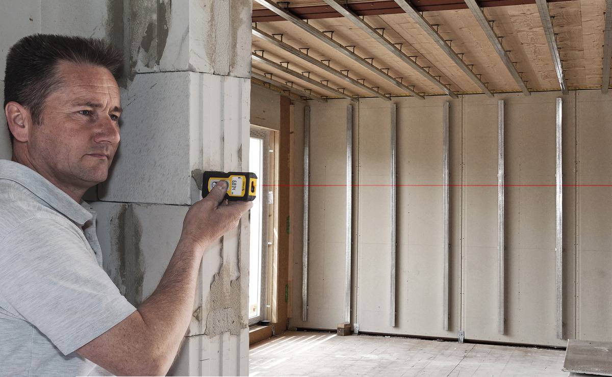 Ein Mann benutzt ein Laser-Entfernungsmessgerät in einem im Bau befindlichen Raum. Eine rote Laserlinie zeigt die Messung an.